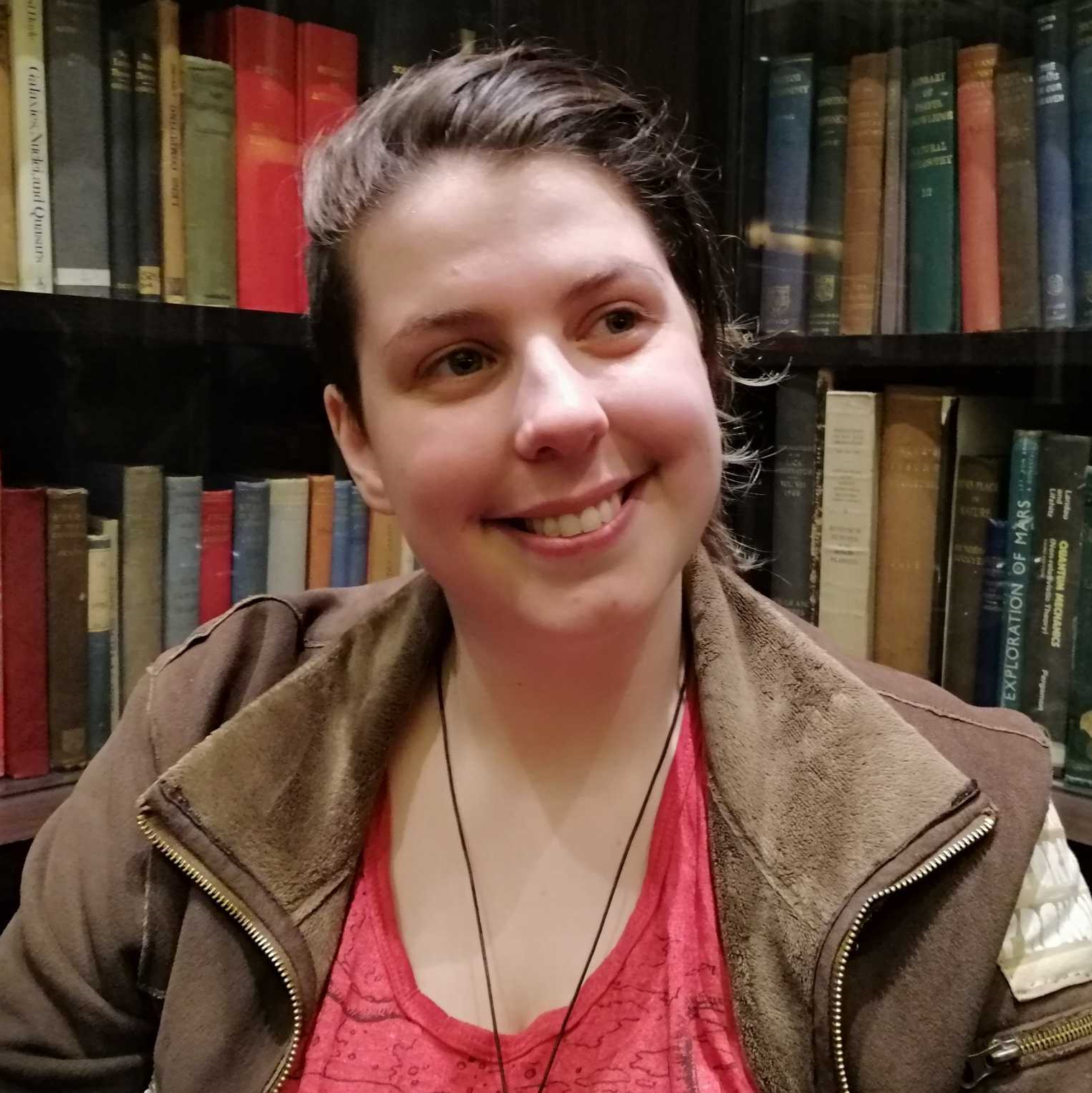 rem is white with brown hair, smiling, wearing a brown suede jacket and in front of shelves of old looking books